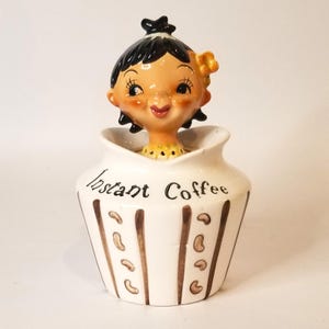 May include: A vintage ceramic instant coffee jar featuring a smiling woman figurine. The white jar has brown vertical stripes and coffee bean designs. The text "Instant Coffee" is written on the jar.