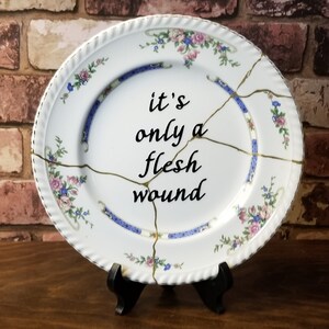 May include: White ceramic plate with floral and blue accents, featuring gold-coloured kintsugi repairs. The plate has the phrase "it's only a flesh wound" in black script. The plate is displayed on a black stand.