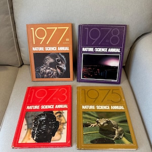 May include: Four vintage "Nature Science Annual" books. The covers are in various colors: orange (1977), purple (1978), red (1973), and gold (1975). Each book features a different nature-themed image, including a bird, a landscape, a sculpture, and a frog.