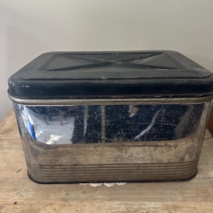 May include: Vintage metal storage container with a black lid and a silver body. The container has a rectangular shape with a ridged design on the lower portion. The lid has a cross-shaped design.