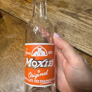 May include: A clear glass Moxie Original soda bottle with an orange and white label. The label features the word "Moxie" in a stylized font, the text "Original", and the phrase "Enjoy a lift...the healthful way."