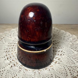 May include: A dark brown, glossy ceramic insulator with a smooth, rounded top section and a wider, cylindrical base. The glaze features subtle mottled variations in tone, and the piece has a distinct, tiered profile with a visible white rim.