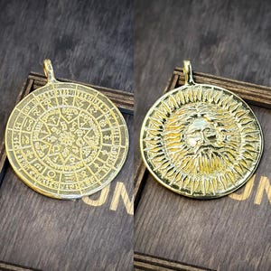 May include: Two gold-coloured pendants. One pendant has an intricate design with symbols and text. The other pendant shows a lion's face surrounded by sun rays. Both pendants have a loop for a chain.