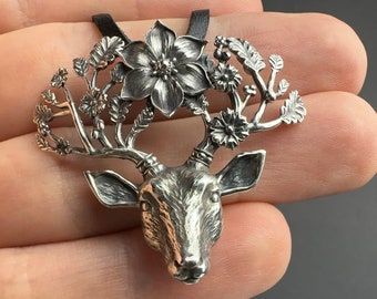 May include: A silver pendant necklace featuring a deer head with antlers adorned with delicate floral details. The antlers are intricately crafted with leaves and flowers, creating a whimsical and nature-inspired design.