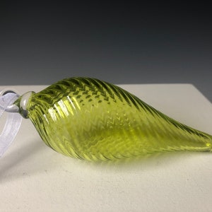May include: A chartreuse glass ornament with a textured, swirled design. The teardrop-shaped ornament has a small loop at the top with a white ribbon for hanging. The ornament is approximately 6 inches long.