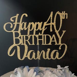 May include: A gold glitter cake topper that reads "Happy 40th Birthday Vania." The topper is placed on a white frosted cake decorated with white daisies and blue flowers. The background is black.