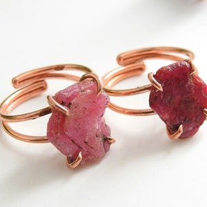 Puede incluir: Dos anillos ajustables de cobre, cada uno con una gema de rubí en bruto. Los rubíes son de un color rojo rosado intenso, y las bandas de cobre tienen un brillo metálico cálido. Los anillos se muestran sobre un fondo blanco.