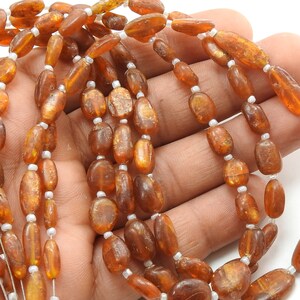 May include: A strand of oval-shaped, amber-colored beads with a golden sheen. The beads are strung on a white thread.