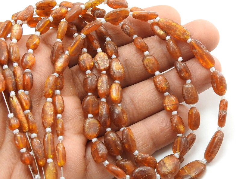 May include: A strand of oval-shaped, amber-colored beads with a golden sheen. The beads are strung on a white thread.