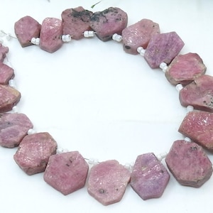 May include: A necklace of rough-cut, pink ruby gemstone beads. The beads are irregularly shaped, with a mix of light and dark pink hues. Each bead is strung on a white cord, creating a necklace-like arrangement. The gemstones have a natural, unpolished appearance.