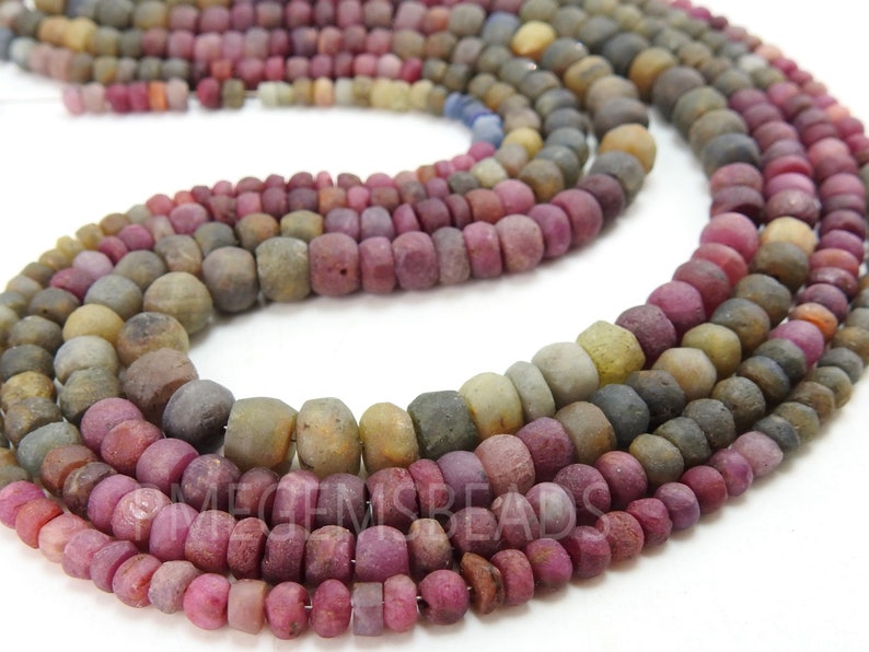 May include: A strand of multi-coloured gemstone beads. The beads are round and have a rough, textured surface. The colours of the beads are shades of pink, red, brown, and grey.