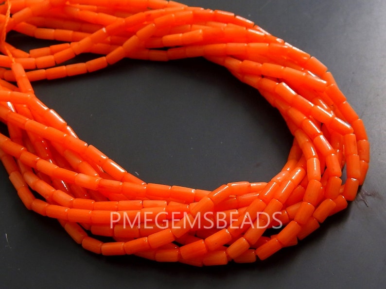 May include: A strand of bright orange cylindrical beads arranged in a circular loop. The beads have a smooth, uniform texture and are strung together on a thin cord. The text PMEGEMSBEADS is printed in white capital letters across the lower section.