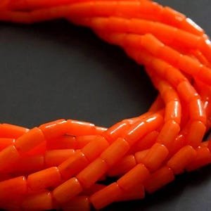 May include: A multi-strand necklace composed of numerous small, cylindrical, bright orange beads. The beads are arranged in a dense, layered formation, creating a continuous, curved loop of vibrant, uniform color throughout the entire piece.
