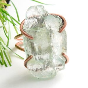 Könnte beinhalten: Ein Ring mit einem großen, rohen, hellgrünen Kristall-Edelstein, der von einer Kupferdrahtfassung gehalten wird. Das Metallband besteht aus einem einzigen Stück Kupferdraht, das sich um den Stein wickelt, um ihn wie eine Kralle zu sichern.
