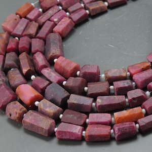 Puede incluir: Primer plano de cuentas de piedras preciosas de rubí facetadas en tonos rojos y burdeos. Las cuentas están ensartadas con pequeños espaciadores blancos. Las piedras tienen una textura natural y rugosa, para joyería.