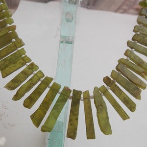 以下が含まれることがあります： 緑色の翡翠のビーズで作られたネックレス。各ビーズは長方形で、わずかに異なる緑色の色合いをしています。ビーズは白い糸に通されています。