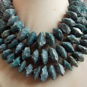 Puede incluir: Un collar de varias hileras hecho con cuentas de piedra preciosa de color verde oscuro, talladas en bruto. Las cuentas tienen una forma irregular y un aspecto natural y rústico.