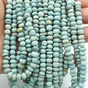 Puede incluir: Una colección de cuentas de color azul claro, redondas y aplanadas, dispuestas en múltiples hilos verticales. Cada cuenta presenta una superficie mate y texturizada con sutiles variaciones de color, incluyendo tonos de verde pálido y beige.