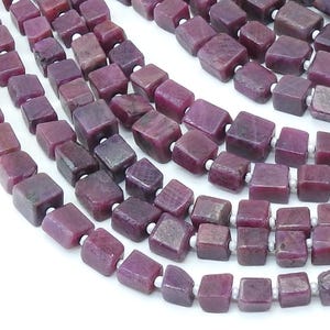 May include: Strands of ruby beads. Each bead is a dark red-purple cube, strung on white thread. The beads have a slightly rough, natural texture. The background is white, highlighting the color and shape of the gemstones.