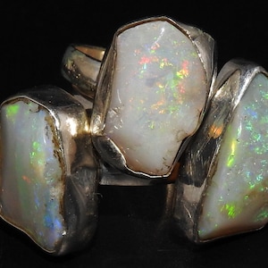 Op de afbeelding: Een zilveren ring met drie grote, witte opaalstenen die in de band zijn gezet. De opalen hebben een regenboogkleurige, iriserende glans.