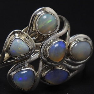 Op de afbeelding: Een zilveren ring met zes opaal edelstenen gezet in een wervelend ontwerp. De opalen zijn iriserend en hebben een verscheidenheid aan kleuren, waaronder wit, blauw en groen.