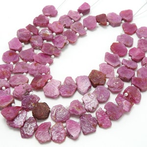 May include: A collection of raw, unpolished pink ruby gemstone slices strung together on thin wire. The stones feature irregular, jagged edges and varying shades of magenta and deep pink, separated by small, round, opaque white spacer beads.