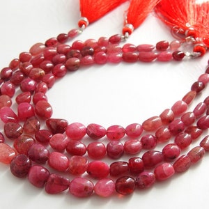May include: A strand of ruby gemstone beads, with a smooth, polished finish. The beads are a deep red color and have a slightly oval shape. The strand is strung on a clear cord and has a tassel at each end.