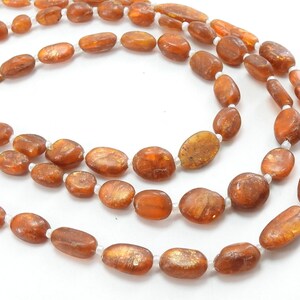 May include: A long strand of brown oval-shaped beads with a shimmery gold finish. The beads are strung on a white thread.