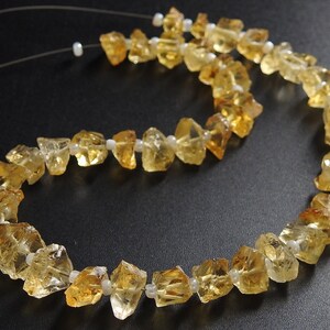 May include: A necklace featuring rough-cut citrine gemstones in shades of yellow and amber. The stones are strung together with small white beads, creating a natural and earthy aesthetic. The necklace is displayed against a dark background.