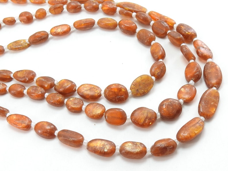 May include: A long strand of brown oval-shaped beads with a shimmery gold finish. The beads are strung on a white thread.