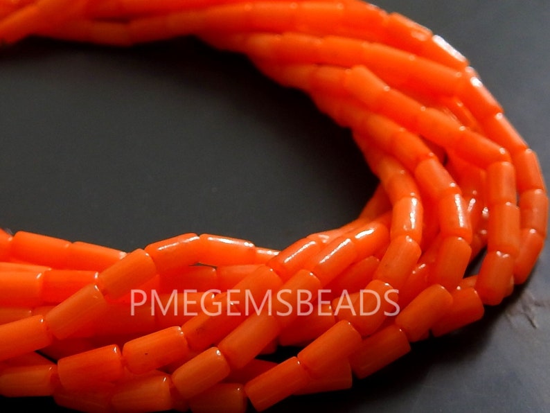 May include: A collection of vibrant orange cylindrical beads arranged in a curved formation. The beads have a smooth, polished finish and a consistent shape. The text PMEGEMSBEADS is clearly visible in white lettering across the center of the beads.