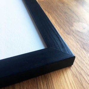 Op de afbeelding: Close-up van een zwart houten fotolijst met een gladde, matte afwerking. De lijst ligt op een houten oppervlak.