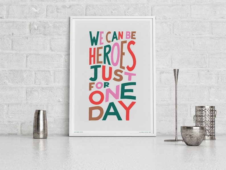 Op de afbeelding: Een ingelijst print met de tekst "WE CAN BE HEROES JUST FOR ONE DAY" in kleurrijke, gestileerde letters op een lichtgrijze achtergrond.