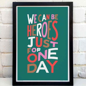 Op de afbeelding: Een turquoise poster met de tekst "WE CAN BE HEROES JUST FOR ONE DAY" in een kleurrijk, gelaagd lettertype. De woorden "HEROES" en "ONE" zijn in roze, "JUST" is in bruin, "FOR" is in oranje, "WE" en "DAY" zijn in rood, en "CAN" en "BE" zijn in wit.