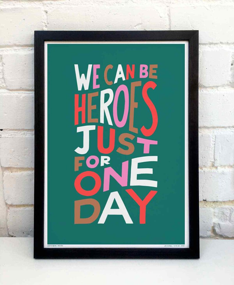 Op de afbeelding: Een turquoise poster met de tekst "WE CAN BE HEROES JUST FOR ONE DAY" in een kleurrijk, gelaagd lettertype. De woorden "HEROES" en "ONE" zijn in roze, "JUST" is in bruin, "FOR" is in oranje, "WE" en "DAY" zijn in rood, en "CAN" en "BE" zijn in wit.