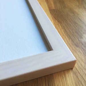 May include: Close-up of the corner of a white wood picture frame. The frame is on a light brown wood surface.