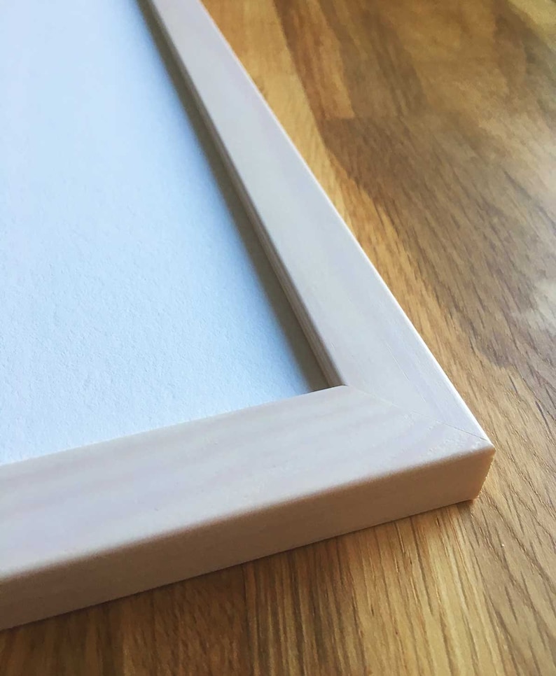 May include: Close-up of the corner of a white wood picture frame. The frame is on a light brown wood surface.
