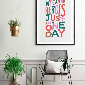 Op de afbeelding: Een ingelijst print met de tekst "We can be heroes just for one day" in kleurrijke, gestileerde letters op een lichtgrijze achtergrond. De print hangt aan een witte muur met een hangende plant en een stoel op de voorgrond.