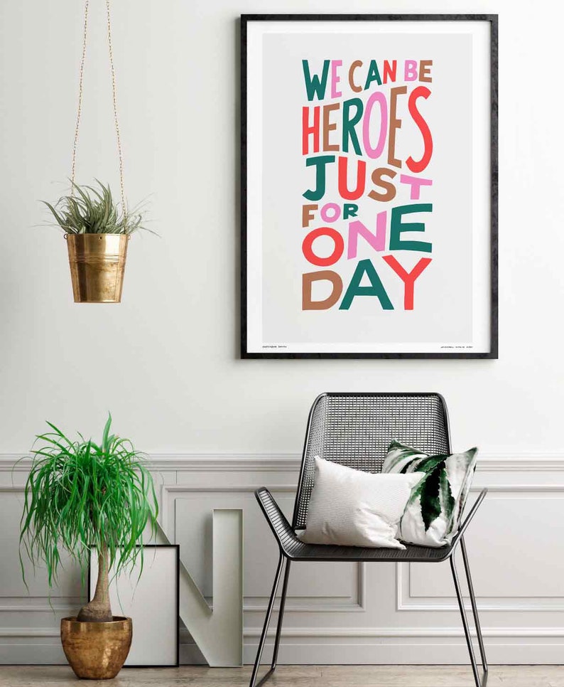 Op de afbeelding: Een ingelijst print met de tekst "We can be heroes just for one day" in kleurrijke, gestileerde letters op een lichtgrijze achtergrond. De print hangt aan een witte muur met een hangende plant en een stoel op de voorgrond.