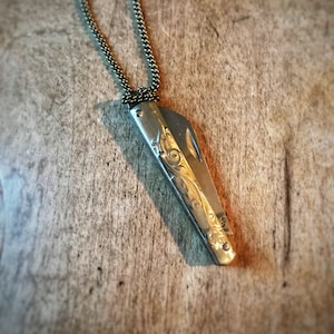 May include: Silver-toned pocket knife pendant with ornate floral engraving, suspended from a matching chain. The knife blade is partially open, revealing a glimpse of the interior. The necklace rests on a wooden surface.