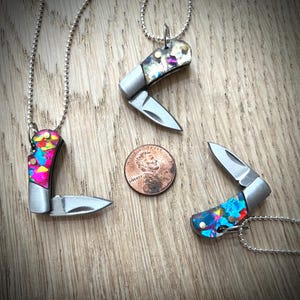 Könnte beinhalten: Drei Miniatur-Taschenmesser-Anhänger mit bunten, gemusterten Griffen und silbernen Klingen. Ein Cent-Stück liegt neben den Messern zum Größenvergleich. Die Messer liegen auf einer Holzoberfläche.