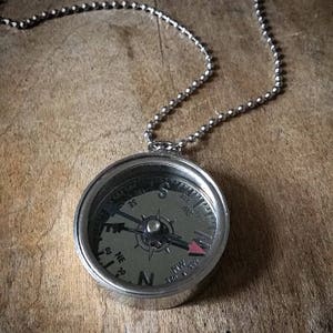 Puede incluir: Un collar de plata con un compás redondo y una cadena de plata. La cara del compás tiene marcas negras y una aguja roja que apunta al norte.