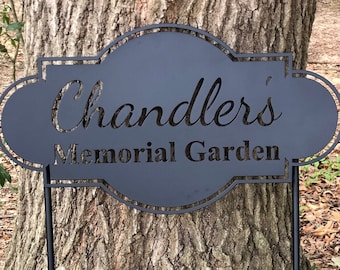 Puede incluir: Señal de metal negro con un borde decorativo y el texto "Chandler's Memorial Garden". La señal está unida a una estaca de metal negro.
