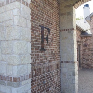 Pu&ograve; includere: Un muro di mattoni con una grande lettera "F" in metallo attaccata ad esso. Il muro &egrave; fatto di pietra chiara e mattoni rosso scuro. La lettera &egrave; nera e ha una finitura rustica.