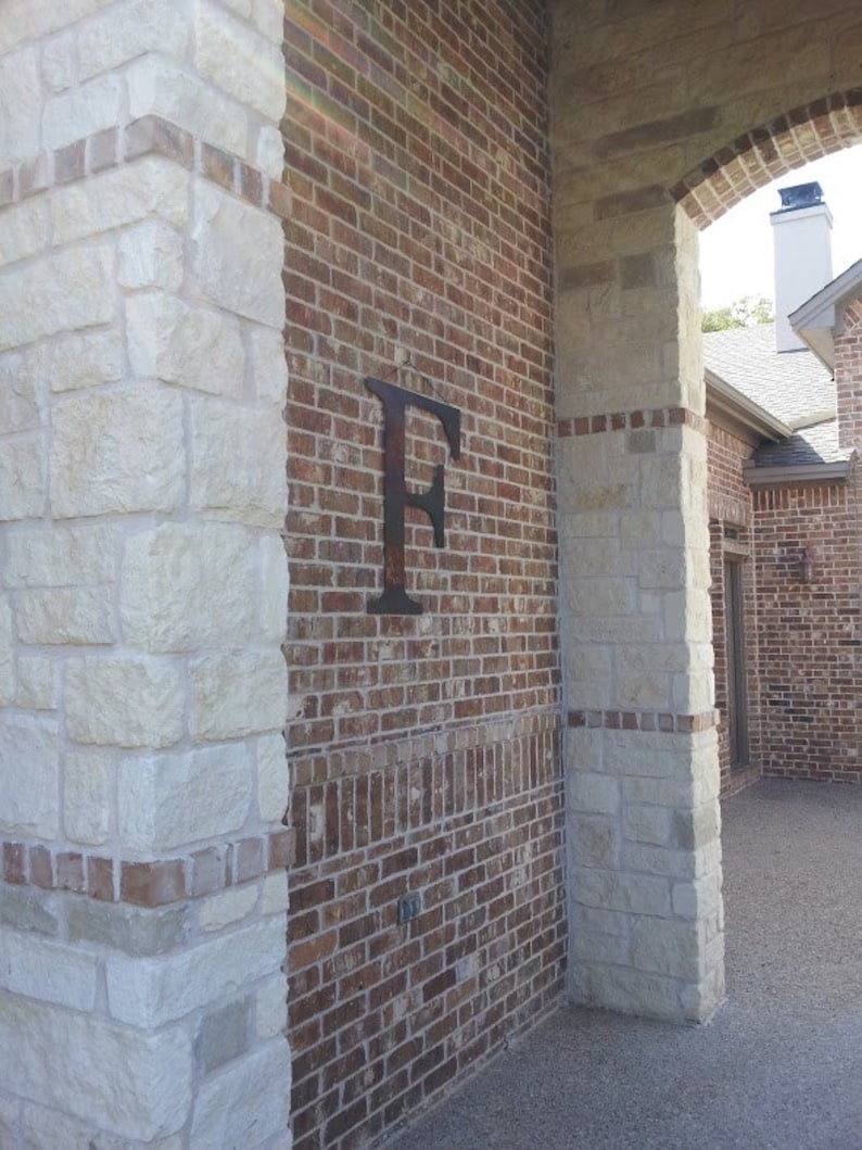 Pu&ograve; includere: Un muro di mattoni con una grande lettera "F" in metallo attaccata ad esso. Il muro &egrave; fatto di pietra chiara e mattoni rosso scuro. La lettera &egrave; nera e ha una finitura rustica.