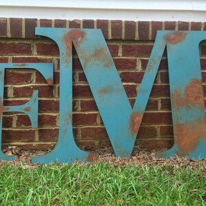 Pu&ograve; includere: Tre grandi lettere metalliche, "E", "M" e "V", dipinte di un colore blu-verde con accenti di ruggine. Le lettere sono in piedi davanti a un muro di mattoni e erba.