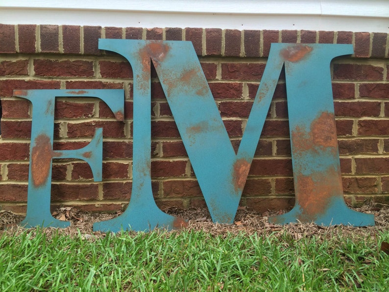 Pu&ograve; includere: Tre grandi lettere metalliche, "E", "M" e "V", dipinte di un colore blu-verde con accenti di ruggine. Le lettere sono in piedi davanti a un muro di mattoni e erba.
