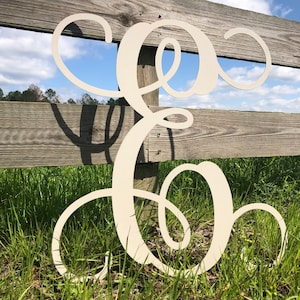 May include: A large, white, wooden letter E with a cursive script font. The letter is leaning against a wooden fence and is surrounded by green grass.