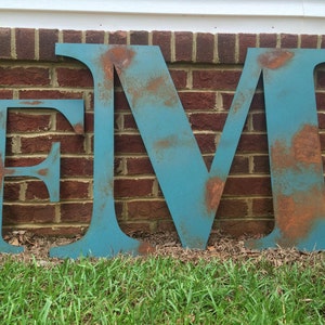 Può includere: Tre grandi lettere metalliche, "E", "M" e "V", dipinte in blu turchese con accenti di ruggine. Le lettere sono in piedi davanti a un muro di mattoni e erba.
