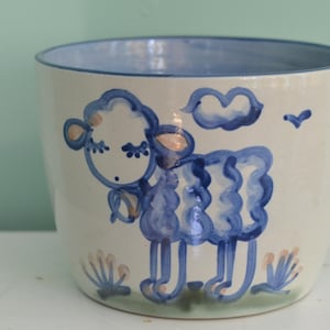 May include: A ceramic planter with a cream-colored base and a hand-painted blue sheep design. The sheep features curly wool, closed eyes, and a small bell around its neck. Above the sheep is a blue cloud, and below are small floral accents.